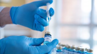 Blue-gloved hands use a needle to extract liquid from a vial.