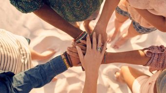 Aerial view of five woman joining their hands together in a semi-circle on the beach.