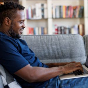 African American male types on a laptop while sitting on a couch.