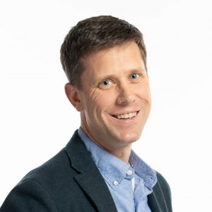 Brunette man in blue shirt and dark jacket smiling.