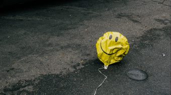 A semi-deflated yellow smiley face balloon lying in a dirty street.