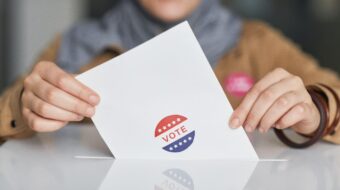 Photo of person dropping a ballot into a box. The person is wearing a hijab.