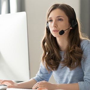 Woman with long brunette hair talking over a headset while looking at a computer monitor.