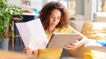 Photo of woman with curly hair looking at tablet and holding a piece of paper