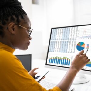 Black woman wearing a mustard yellow shirt and holding a pen and phone while analyzing data charts and graphs on a computer screen