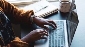 A person typing on a MacBook. They are wearing an Apple Watch and a gold wedding ring set. They have a cup of tea next to them and a notebook and pen.