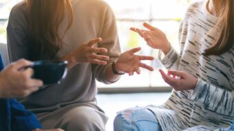 Three people engaged in a lively conversation, using their hands to emphasize their communication. One person is holding a cup of coffee.